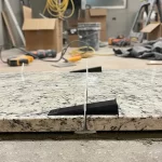 Imagem de um espaçador para mármores e granitos usado para alinhar placas de pedra durante a instalação, com ambiente de oficina ao fundo.