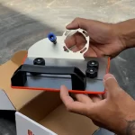 Suporte de esmerilhadeira para corte e chanfro de 45 graus, sendo instalado em uma bancada de trabalho com uma caixa de papelão ao fundo.